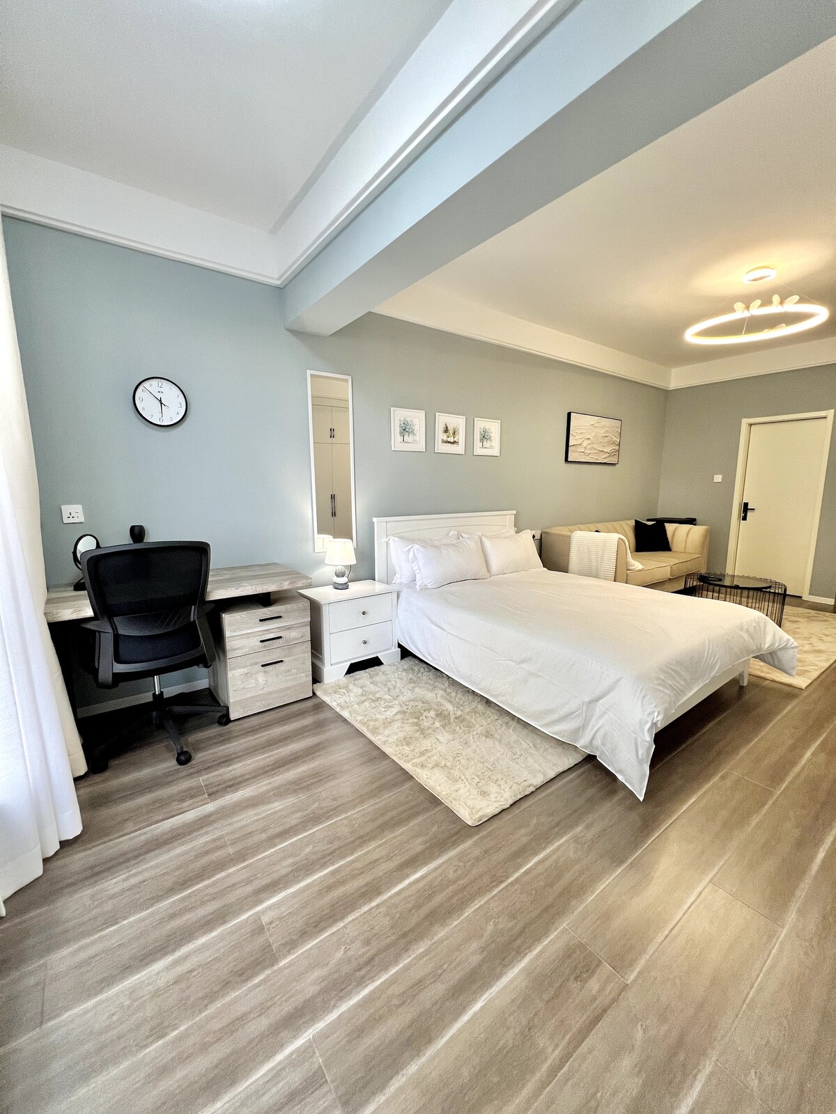 A spacious studio features a comfortable bed with crisp white linens, a stylish desk with an office chair, and light wood flooring. Neutral-toned walls are accented by framed artwork. Natural light enters through sheer curtains, creating a bright and inviting atmosphere.