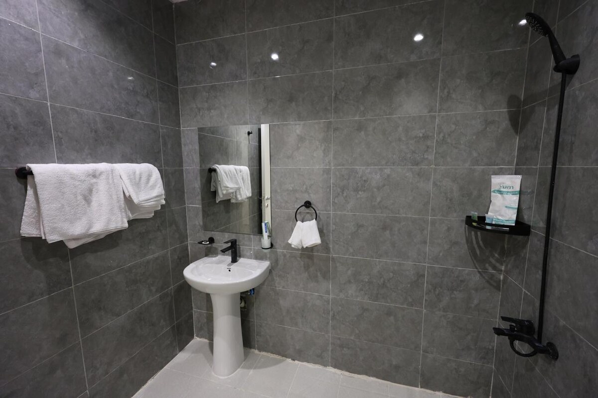 A modern bathroom features slate-grey tiles and bright lighting. A sleek sink is positioned next to a large mirror. Hand towels are neatly hung on the wall, while a soap dispenser and shower accessories are arranged on a wall-mounted shelf. A spacious shower area is included.