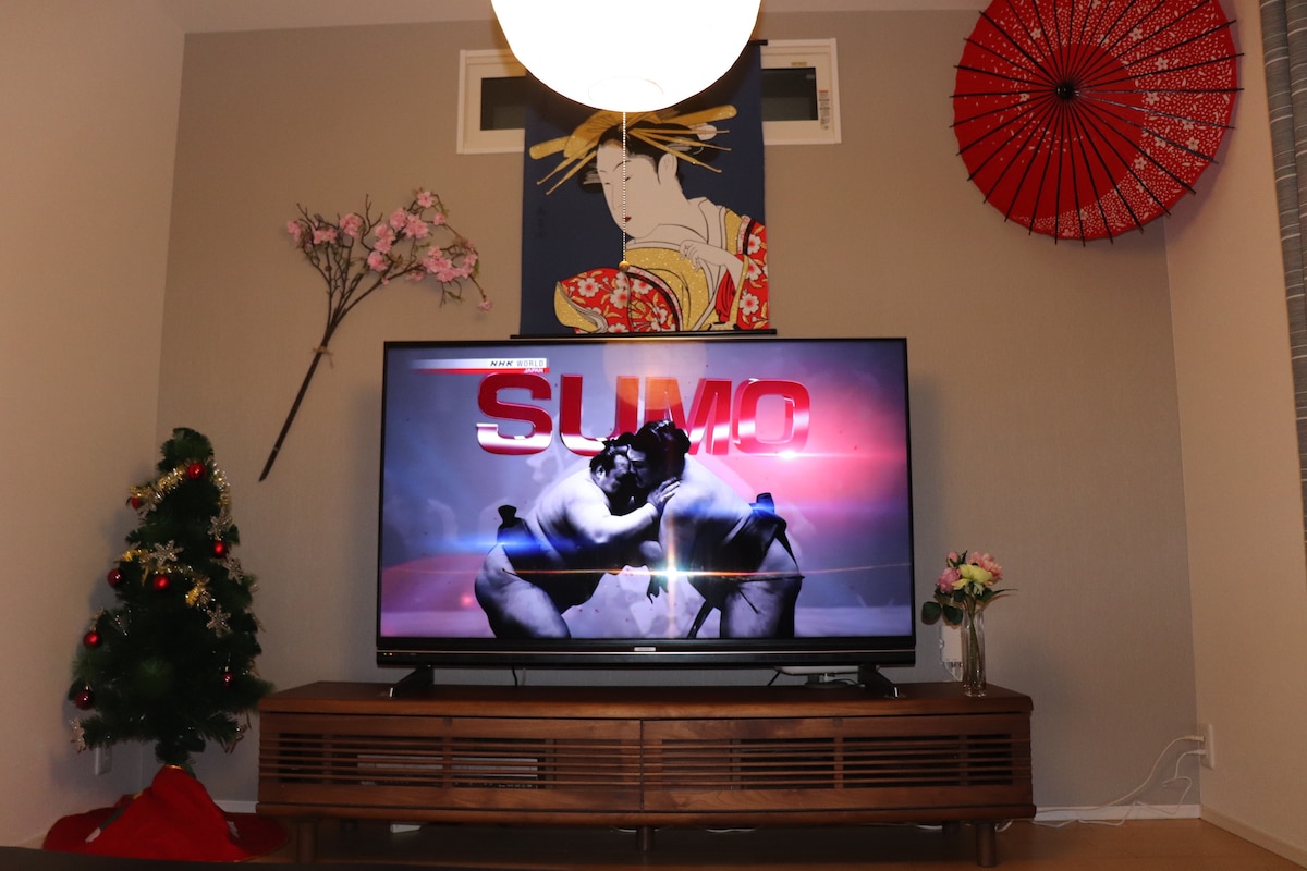 A large television is positioned on a wooden media console, displaying a sumo wrestling match. A colorful wall art piece featuring a woman in traditional attire hangs above the TV. Decorative elements include a small Christmas tree, a floral arrangement, and a red umbrella mounted on the wall.