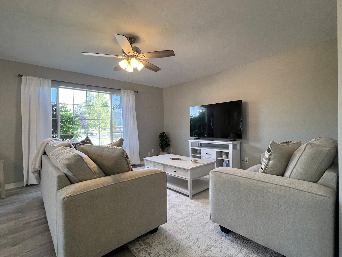 A light-filled living area features a large window with sheer white curtains. Two plush sofas face a white coffee table, while a flat-screen television is mounted on the wall. A ceiling fan offers comfort, and neutral tones create a relaxing environment.