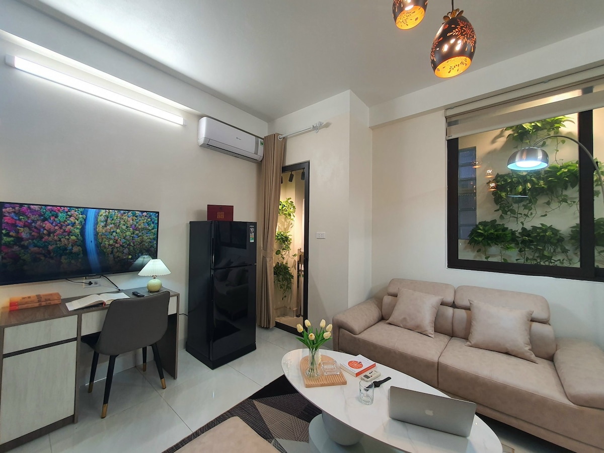 A modern living area features a comfortable beige sofa, a stylish coffee table, and a desk with a chair. A large TV displays colorful imagery, while a fridge is positioned nearby. Natural light filters through the window, highlighting indoor plants.