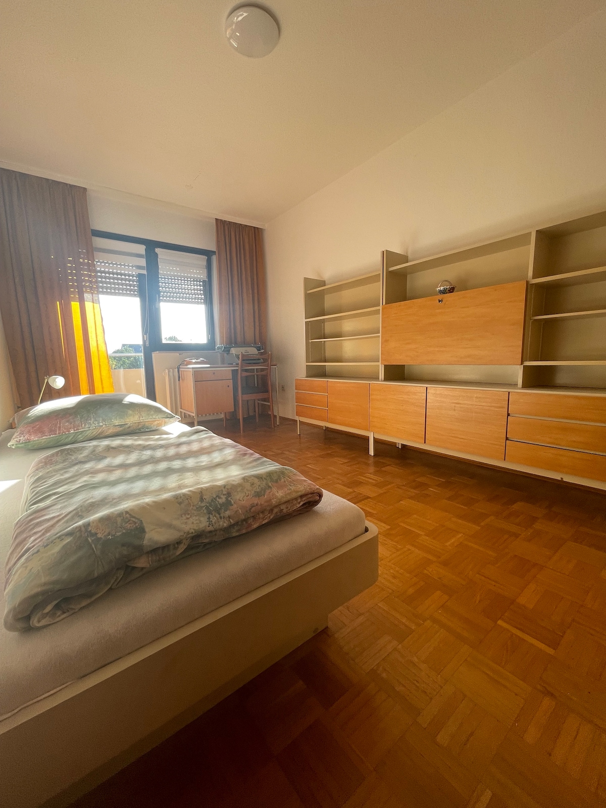 A cozy bedroom features a spacious bed with patterned bedding. Light streams in through a window, enhancing the warm wooden floors. A modern wooden shelf unit and cabinet offer storage space, while sheer curtains add a touch of softness to the room.