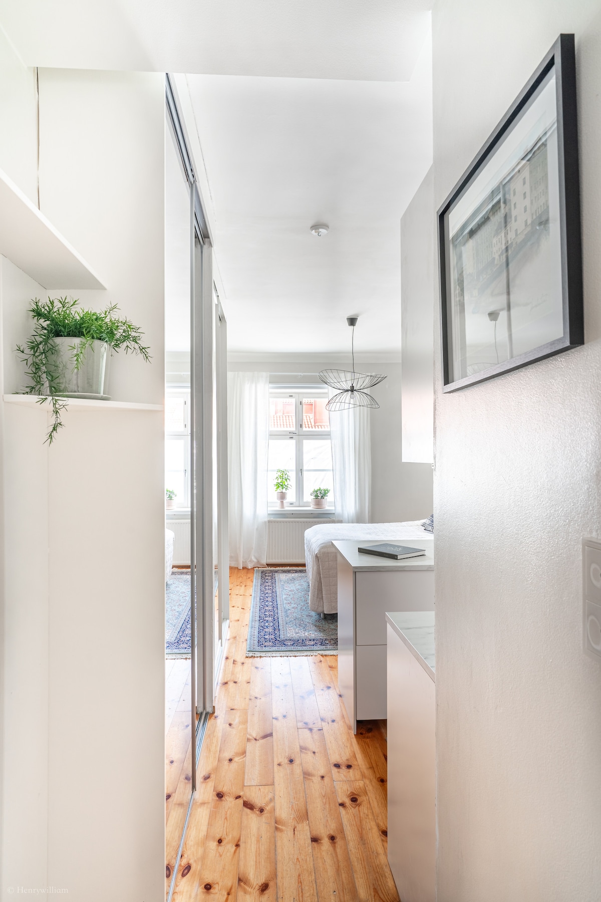 A narrow hallway leads to a bright bedroom with large windows, allowing natural light to fill the space. Light wood flooring adds warmth, and a decorative rug enhances the inviting atmosphere. Green plants on shelves provide a touch of greenery, complemented by wall art.