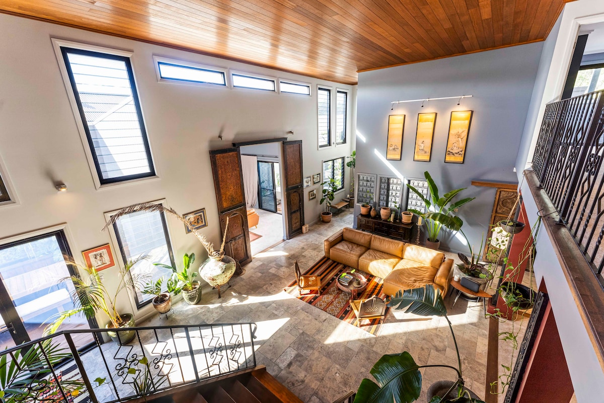 A spacious living area features high ceilings and large windows, allowing natural light to fill the space. Authentic antique Moroccan doors and decorative plants add character. A comfortable leather sofa sits atop a patterned rug, complemented by artwork and a warm stone floor.