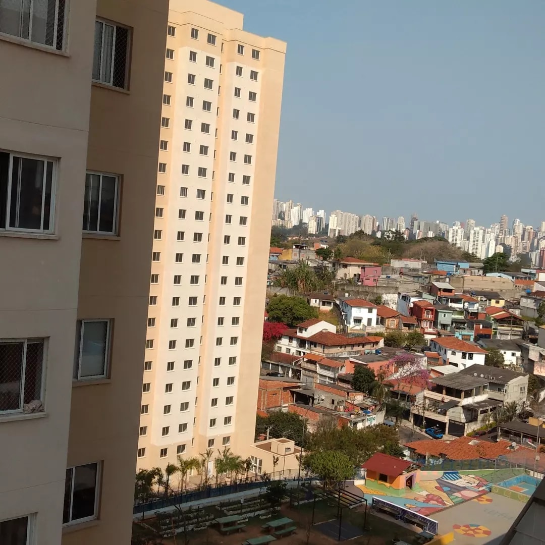 The image displays a view from a high vantage point, featuring a cityscape with a mix of contemporary buildings and colorful residential houses. Lush greenery is visible in the foreground, while a playground area is observed below. The skyline includes numerous high-rise structures in the distance.
