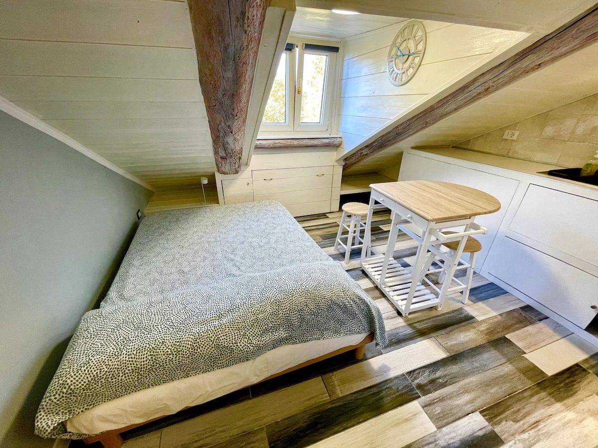 The intimate sleeping area features a spacious bed with a patterned blanket, complemented by light wood flooring. A small, round table with stools is positioned nearby, and natural light enters through windows beneath sloped ceilings, enhancing the cozy atmosphere.