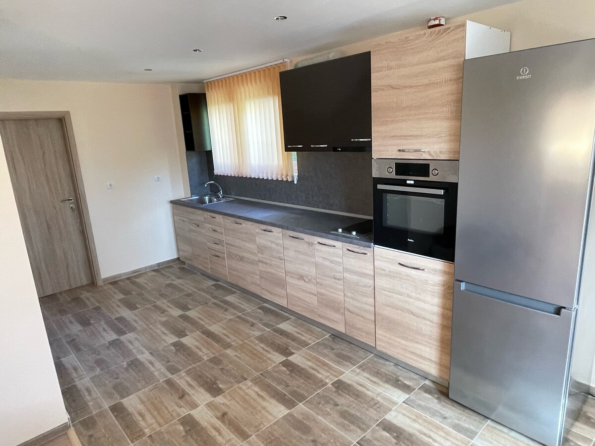 A modern kitchen is presented with wood-toned cabinetry and sleek countertops. Stainless steel appliances include an oven and refrigerator. Natural light filters through cream-colored curtains, illuminating the spacious area with tiled flooring.