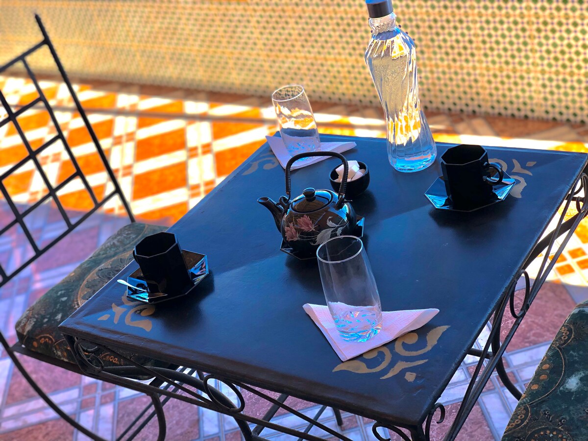 A traditional Moroccan table is set with a black teapot, two black cups, and two glasses. Elegant patterns adorn the table's surface, complemented by a decorative tablecloth. The backdrop features geometric tile designs, enhancing the cultural ambiance.