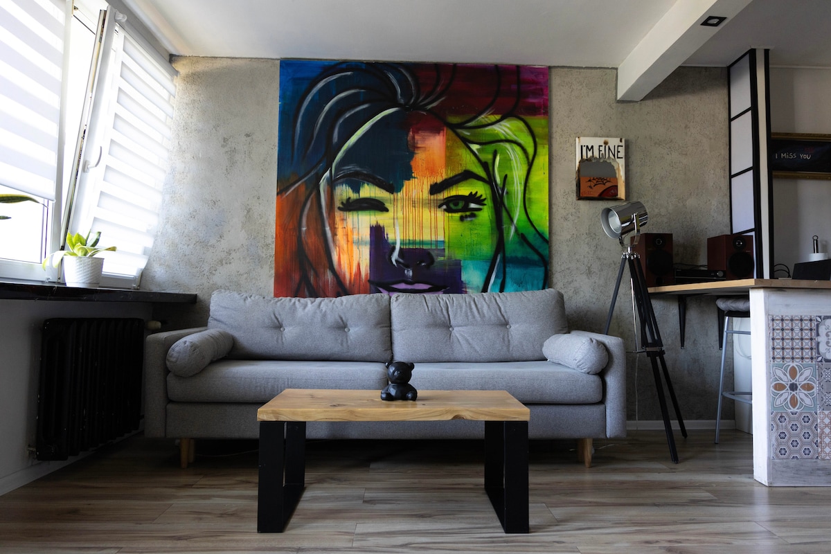 A modern living area features a gray sofa centered against a textured wall, adorned with a vibrant, large-scale artwork of a face. A wooden coffee table sits in front, accompanied by a small decorative figure. Subtle natural light enters through sleek window blinds.