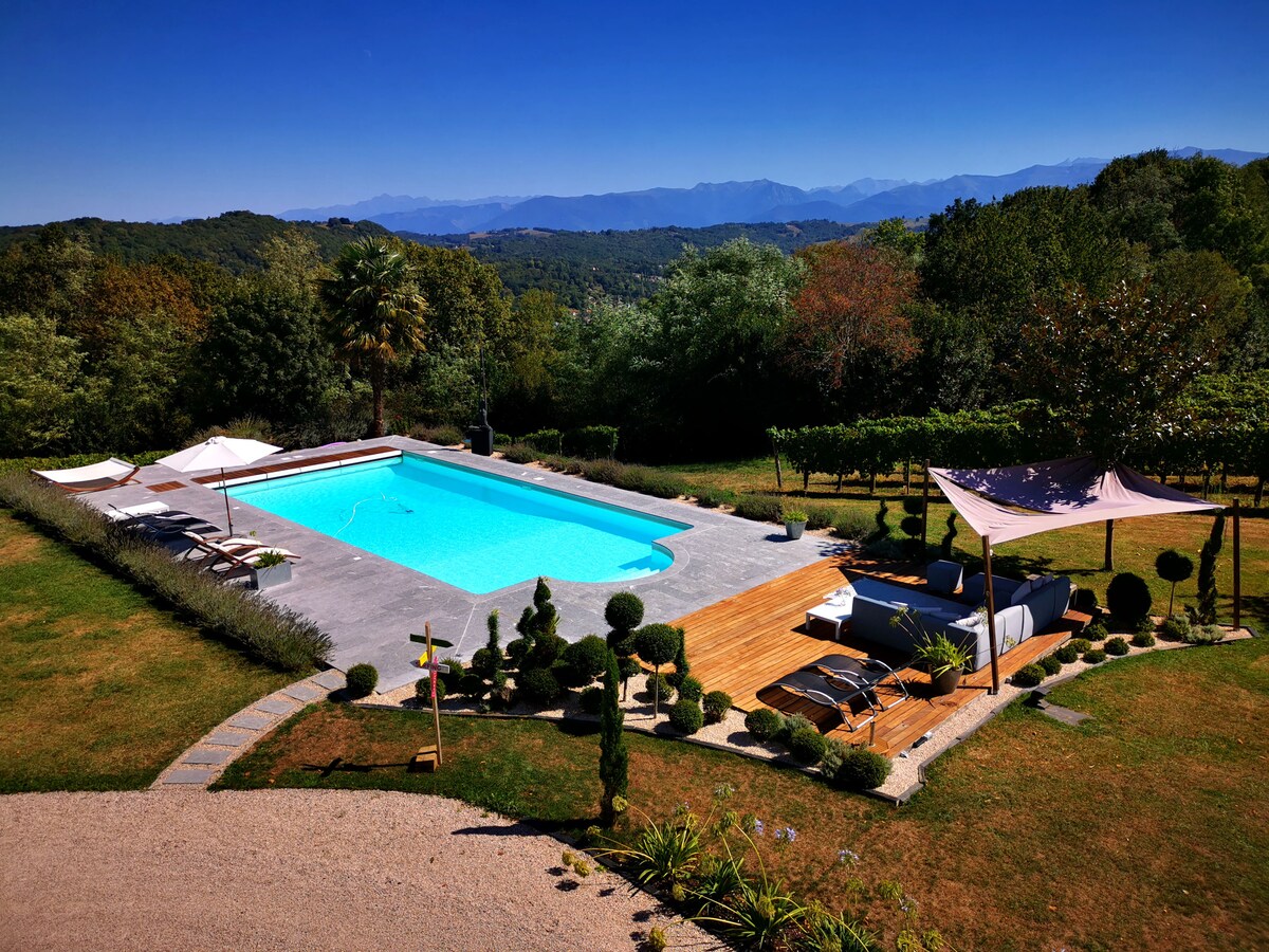 An expansive outdoor area features a refreshing blue swimming pool surrounded by lounge chairs and shaded umbrellas. Lush greenery and neatly trimmed hedges provide a serene atmosphere, while distant mountains create a picturesque backdrop, enhancing the feeling of tranquility and nature.