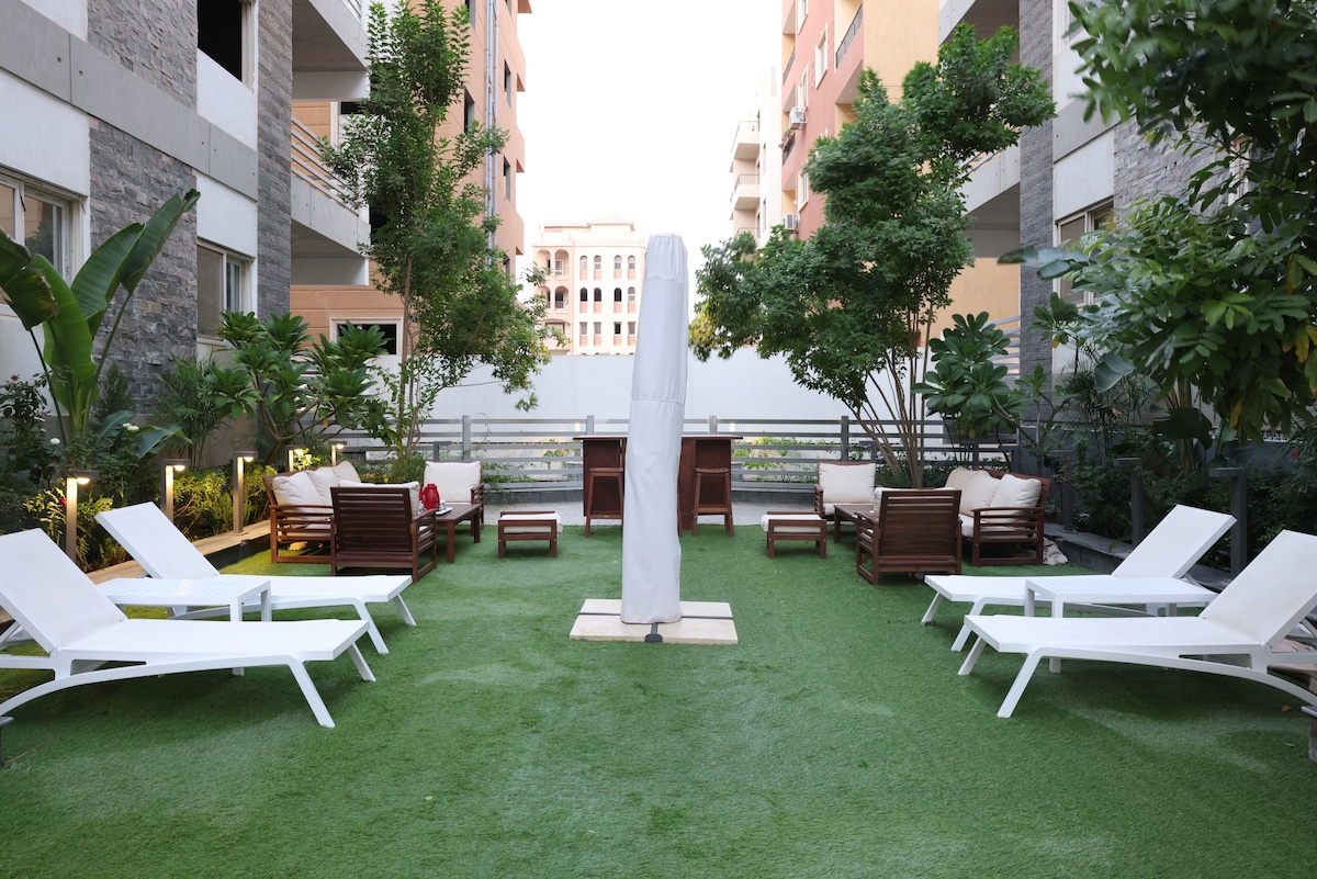 A landscaped garden area features several lounge chairs arranged on green grass, with trees and plants providing a natural ambiance. A shaded structure stands in the center, creating a space for relaxation and social activities.