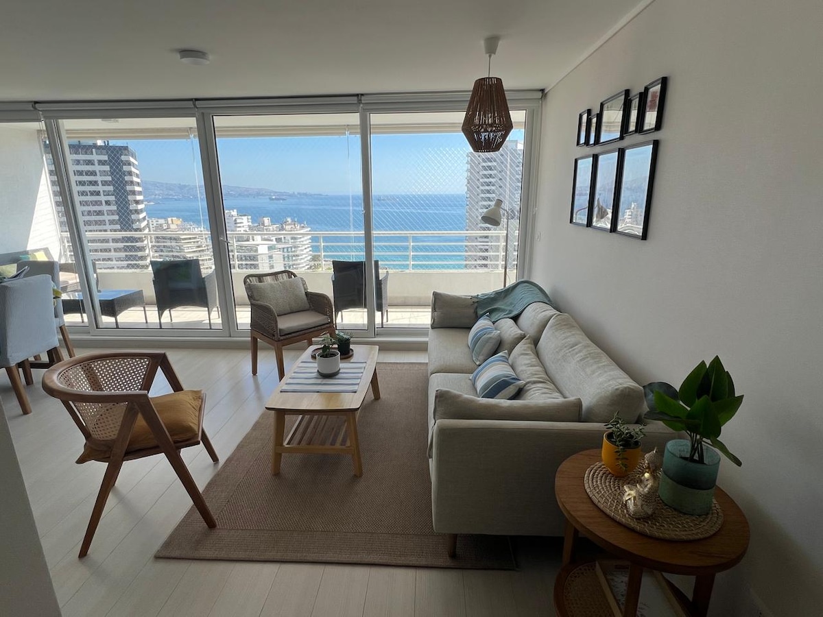 A modern living area is highlighted by a cozy sofa and a wooden coffee table, surrounded by two chairs. Large windows provide a view of the ocean and city skyline, while light enters through the blinds. Decorative plants and wall art add a touch of charm.