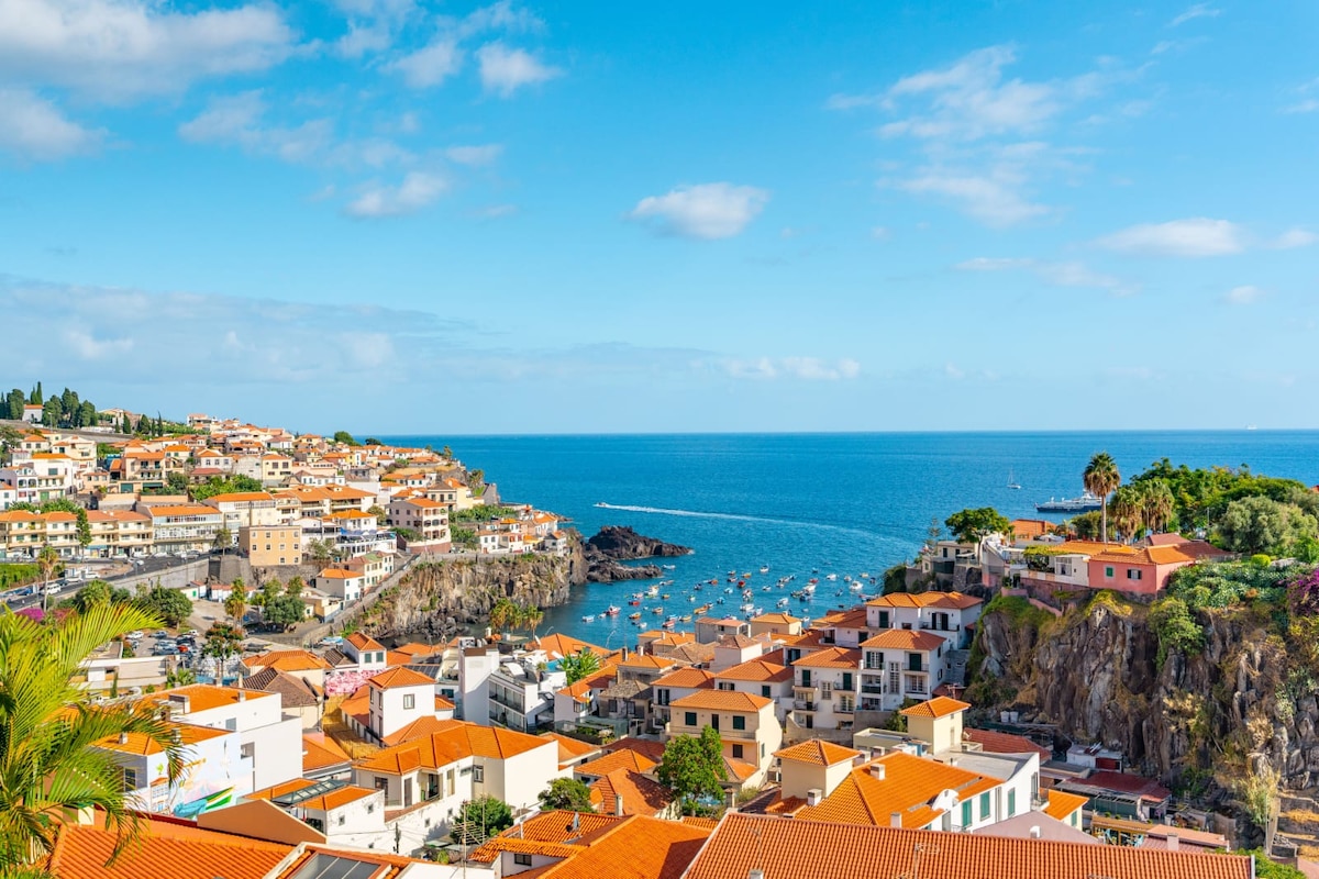 A panoramic view captures the vibrant rooftops of Câmara de Lobos, adorned with orange tiles, overlooking the azure sea. Small boats are visible in the harbor, complemented by the lush greenery on the coastline. The clear sky enhances the scenic beauty of the coastal landscape.