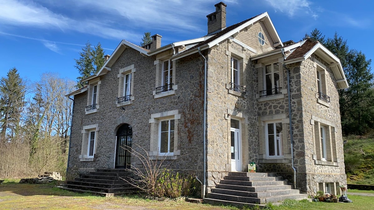 A historic villa features a grey stone exterior with large windows and balconies. The entrance is framed by stairs leading up to the front door. Surrounding greenery is visible, creating a natural setting for the home. A clear blue sky adds brightness to the scene.