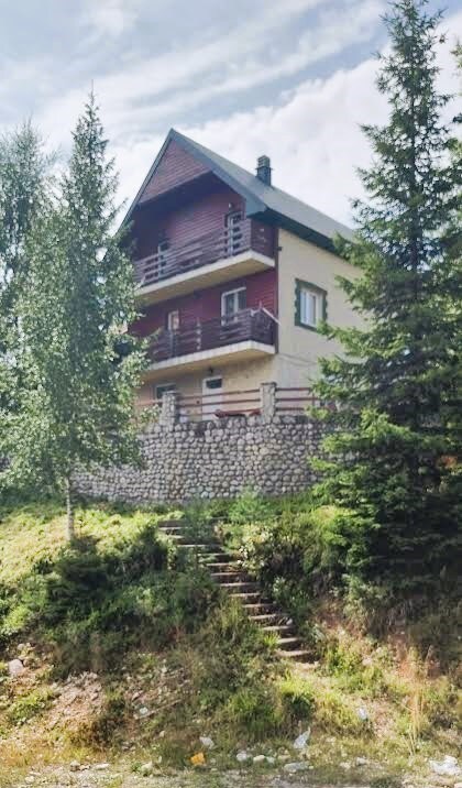 A three-story house is surrounded by trees, with a stone base and wooden accents. The exterior features balconies and a sloped roof, providing a charming architectural style. A set of steps leads from the greenery to the entrance of the home.