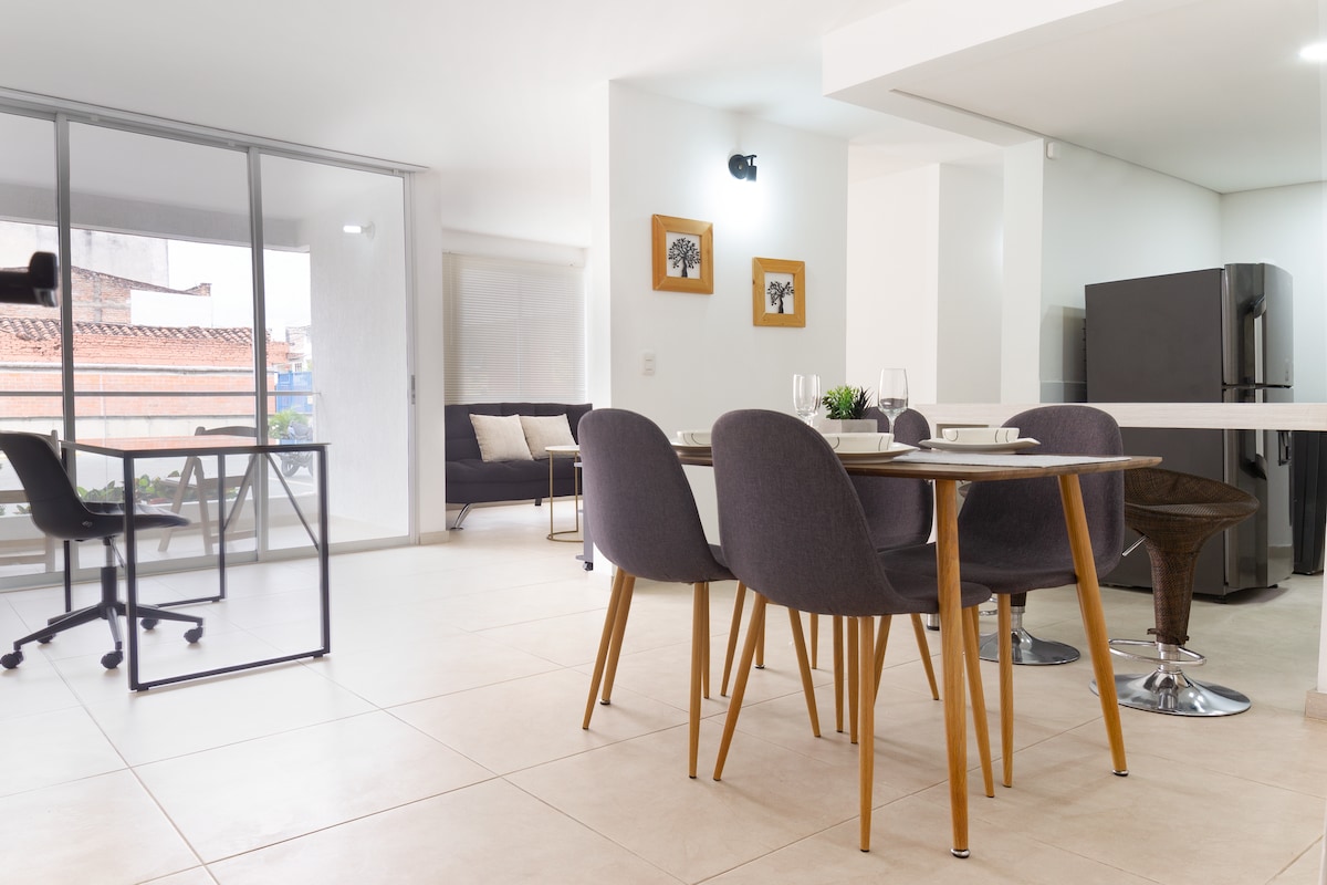 An open-plan living space features a dining table with four chairs, complemented by a sleek gray sofa. A large window allows natural light to fill the room, while a modern kitchen and a dark refrigerator are visible in the background. A workspace chair is positioned nearby.