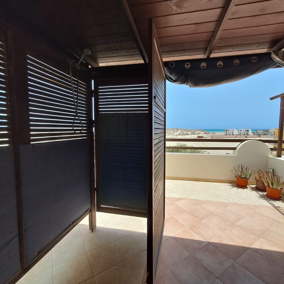 An outdoor shower area is enclosed by wooden slats, creating a sense of privacy. The sea is visible in the distance, enhancing the serene atmosphere. Light-colored tiles cover the floor, and potted plants line the edge, contributing to the relaxed setting.