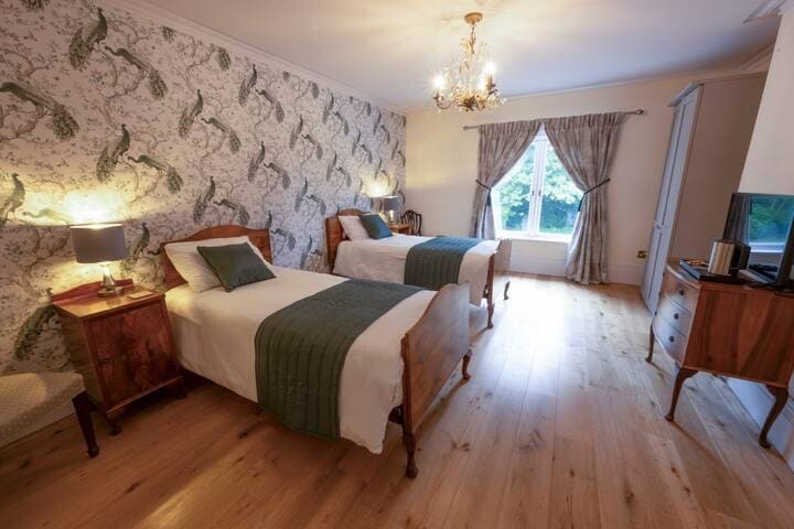 The Nymans room features two single beds dressed in white linens and green accent pillows. A patterned wallpaper with a nature theme complements the light wood flooring. A large window allows natural light to fill the space, and a chandelier adds a classic touch.