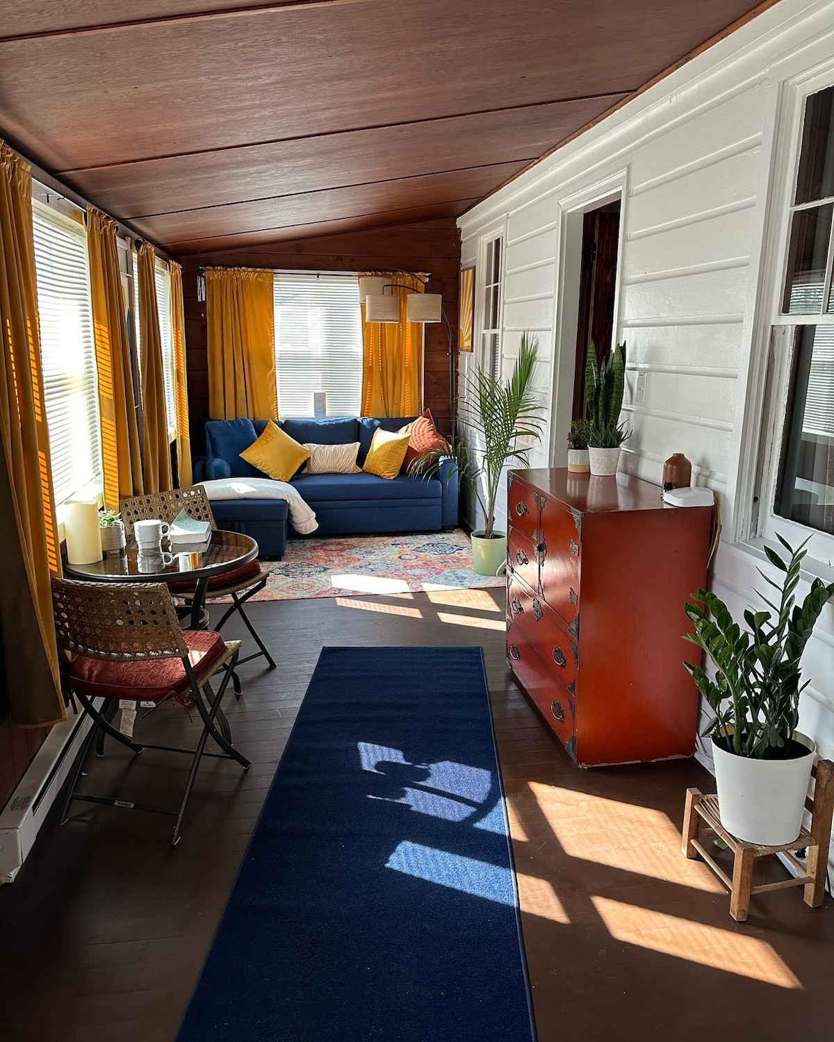 The sunlit sunroom features a bright blue sofa adorned with yellow and white cushions, complemented by a colorful area rug. A small table with chairs is set for coffee, while plants add a touch of nature. Large windows allow ample natural light to fill the space.