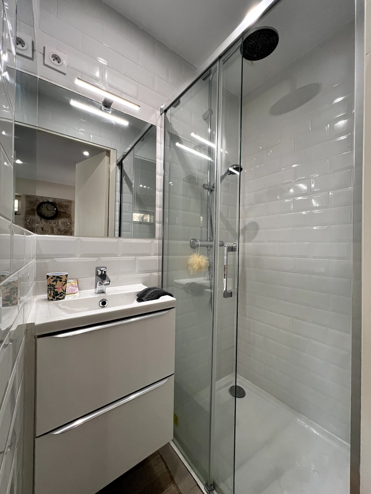 A modern bathroom features a glass-enclosed shower with a rainfall showerhead. White subway tiles cover the walls, and a sleek vanity with storage is visible. Soft lighting illuminates the space, highlighting the minimalist design.