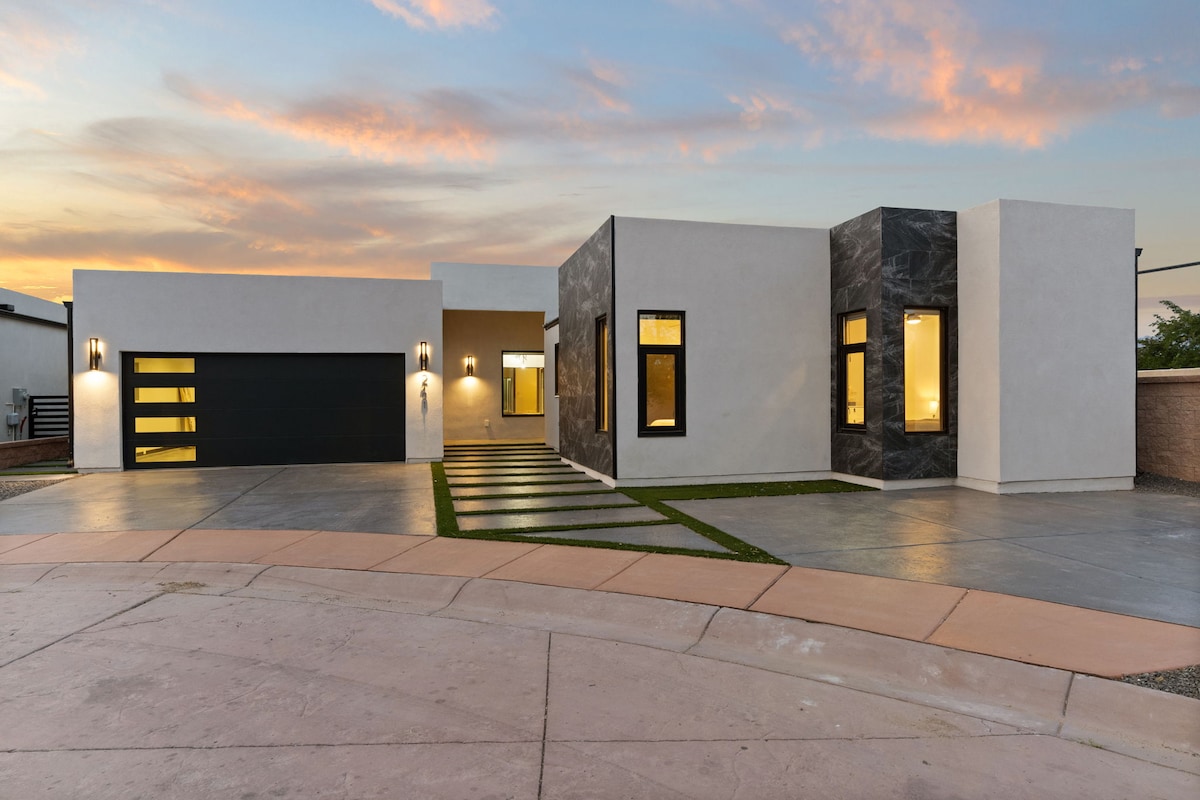 A modern home exterior is presented with a combination of sleek stucco and stone finishes. Large windows allow for ample natural light, while the driveway leads to a garage with a contemporary door design. Soft landscaping and a pathway enhance the property’s curb appeal.