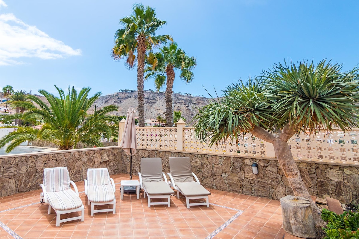 A spacious outdoor terrace is presented, featuring four lounge chairs arranged for relaxation. Lush palm trees and a vibrant greenery surround the area, while a stone wall provides privacy. A clear blue sky enhances the inviting atmosphere of the space.