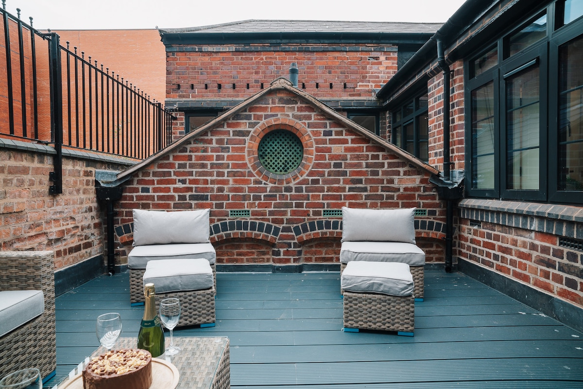 An outdoor terrace features a combination of wicker seating arranged around a small table. Exposed brick walls create a warm backdrop, complemented by large windows that invite natural light. The space is designed for relaxation, perfect for enjoying drinks with friends.
