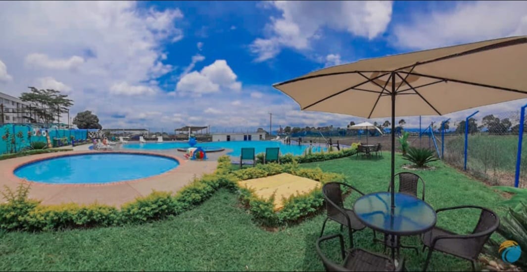 An outdoor area features a pool surrounded by lush greenery, complemented by lounge chairs and umbrellas for shade. A patio table with chairs provides a space for gathering. The sky is partly cloudy, enhancing the inviting atmosphere of the pool area.