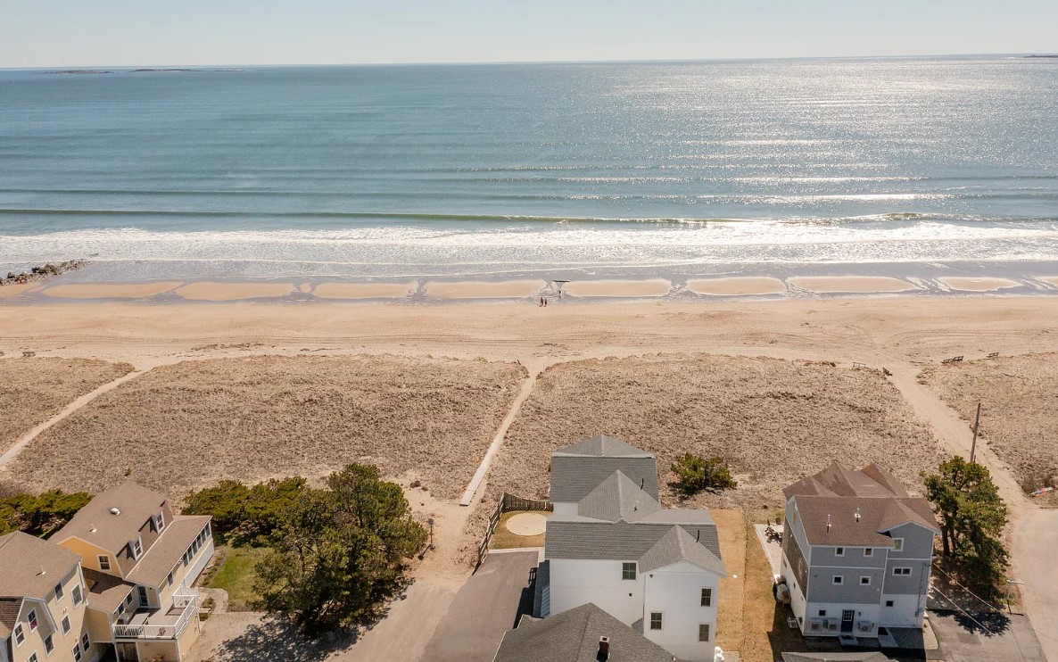 Aerial view captures a serene beach scene with rolling ocean waves meeting a sandy shore. Beachfront properties line the area, while a clear sky and shimmering water enhance the tranquil setting. The image reflects the expansive shoreline, inviting relaxation and leisurely pursuits.