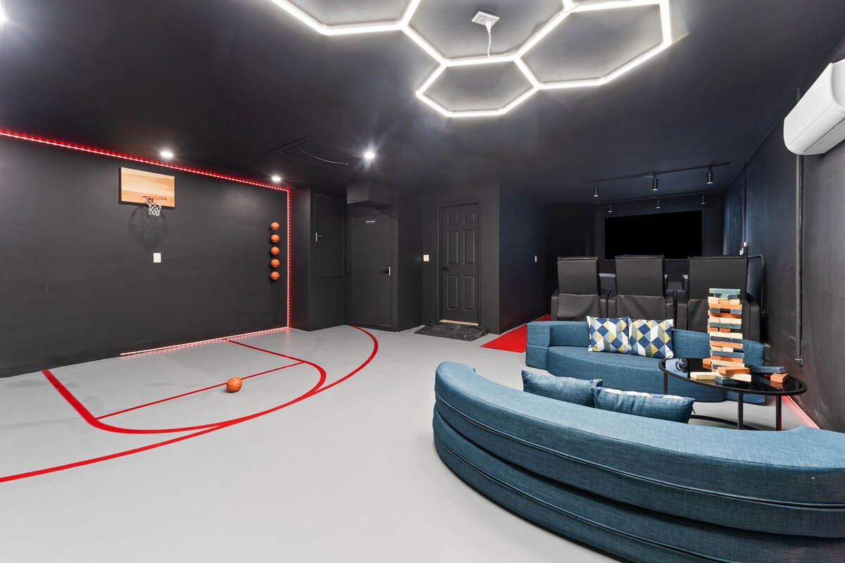 A modern game room showcases a private basketball court with red markings and a hoop. Comfortable seating is provided by a curved blue couch, contrasting against the dark walls. LED lighting highlights the space, creating a lively atmosphere, while a stack of games is visible on the table.