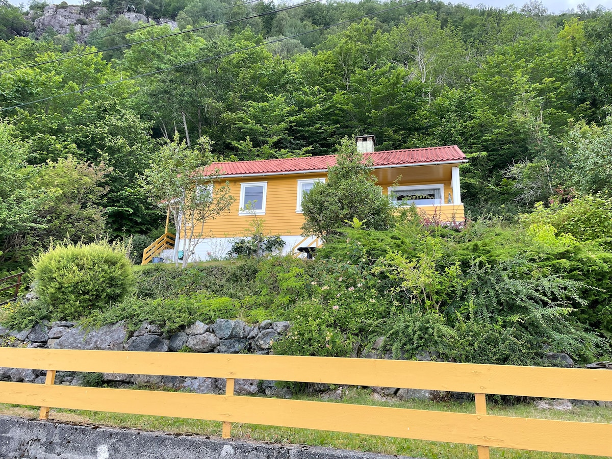 A quaint yellow cabin sits on a hillside, surrounded by lush greenery and trees. A stone wall borders the property, and a wooden staircase leads up to the entrance, enhancing the inviting feel of the natural landscape.