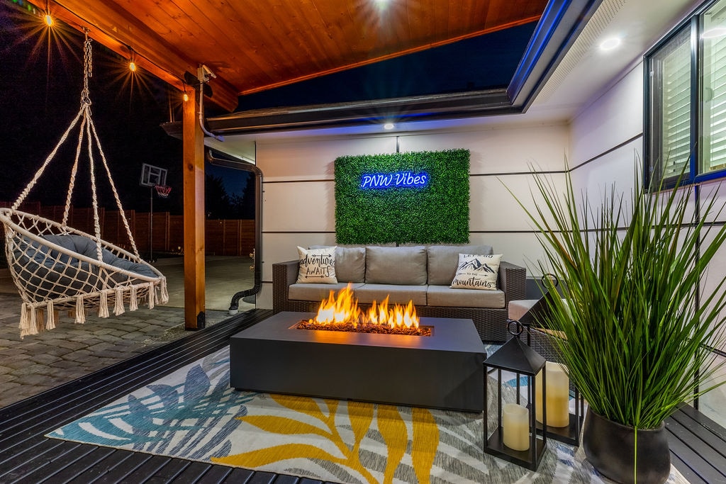 An inviting outdoor space features a modern firepit surrounded by comfortable seating. A swinging chair hangs nearby, complemented by decorative cushions. Soft lighting and greenery enhance the area, which is designed for relaxation and social gatherings in the evening.