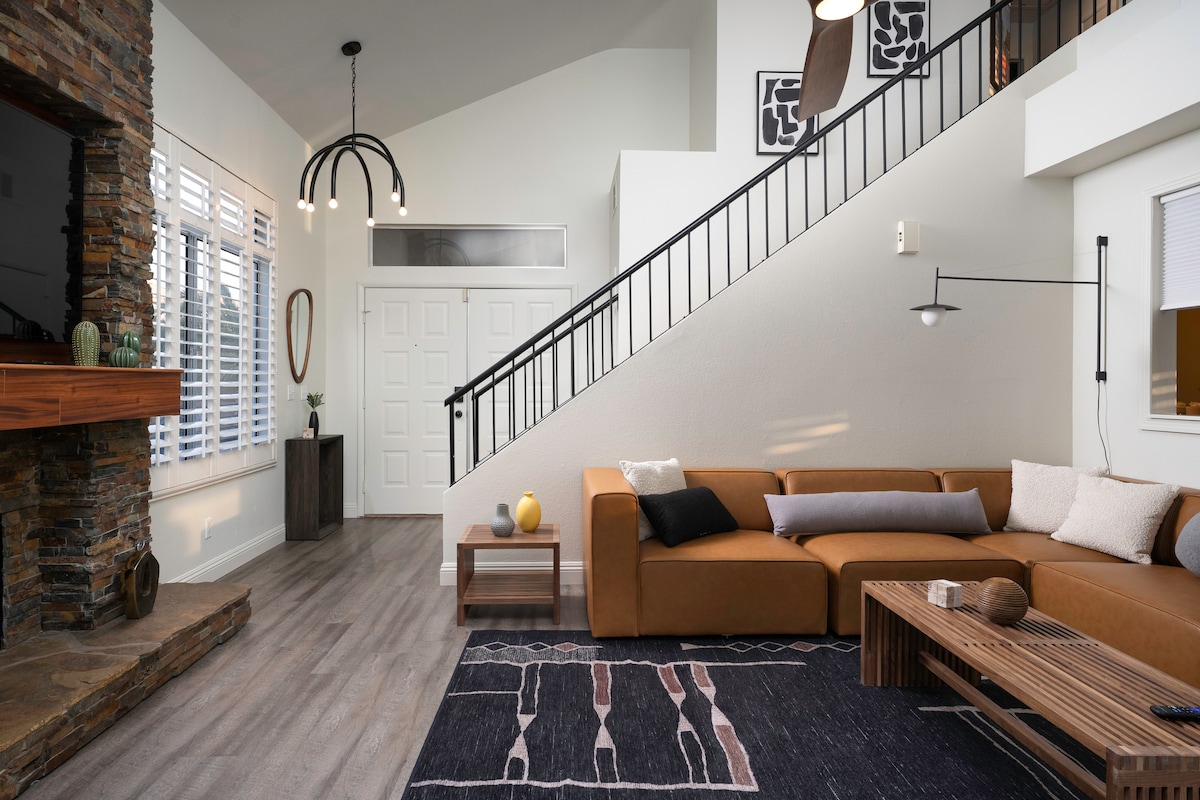A spacious living area is highlighted by a contemporary sectional sofa in earthy tones, complemented by a wooden coffee table. Natural light filters through the grid-patterned windows, illuminating the neutral walls and sleek staircase. A decorative stone fireplace adds warmth to the inviting ambiance.