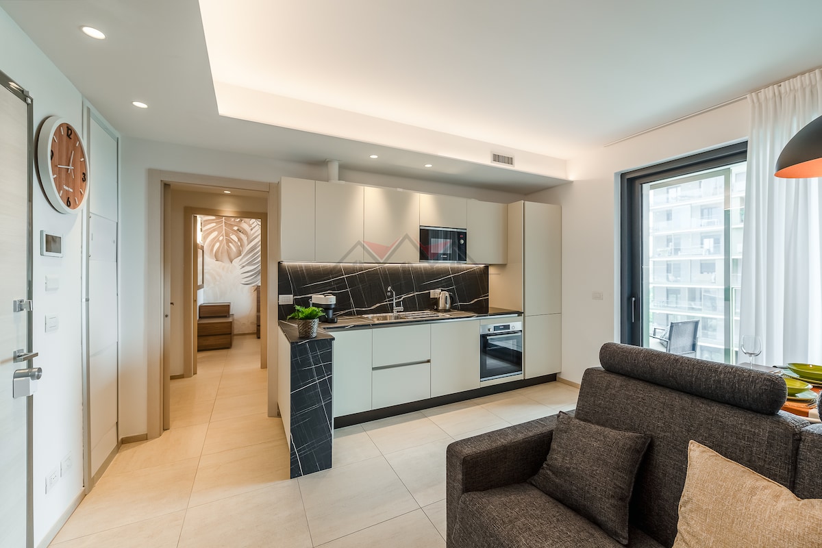A modern kitchen is featured with sleek cabinetry and a black tile backsplash. Included are an induction cooktop, microwave, and refrigerator, complemented by natural light from a nearby window. A cozy seating area is arranged with a gray sofa, providing a warm and inviting space.