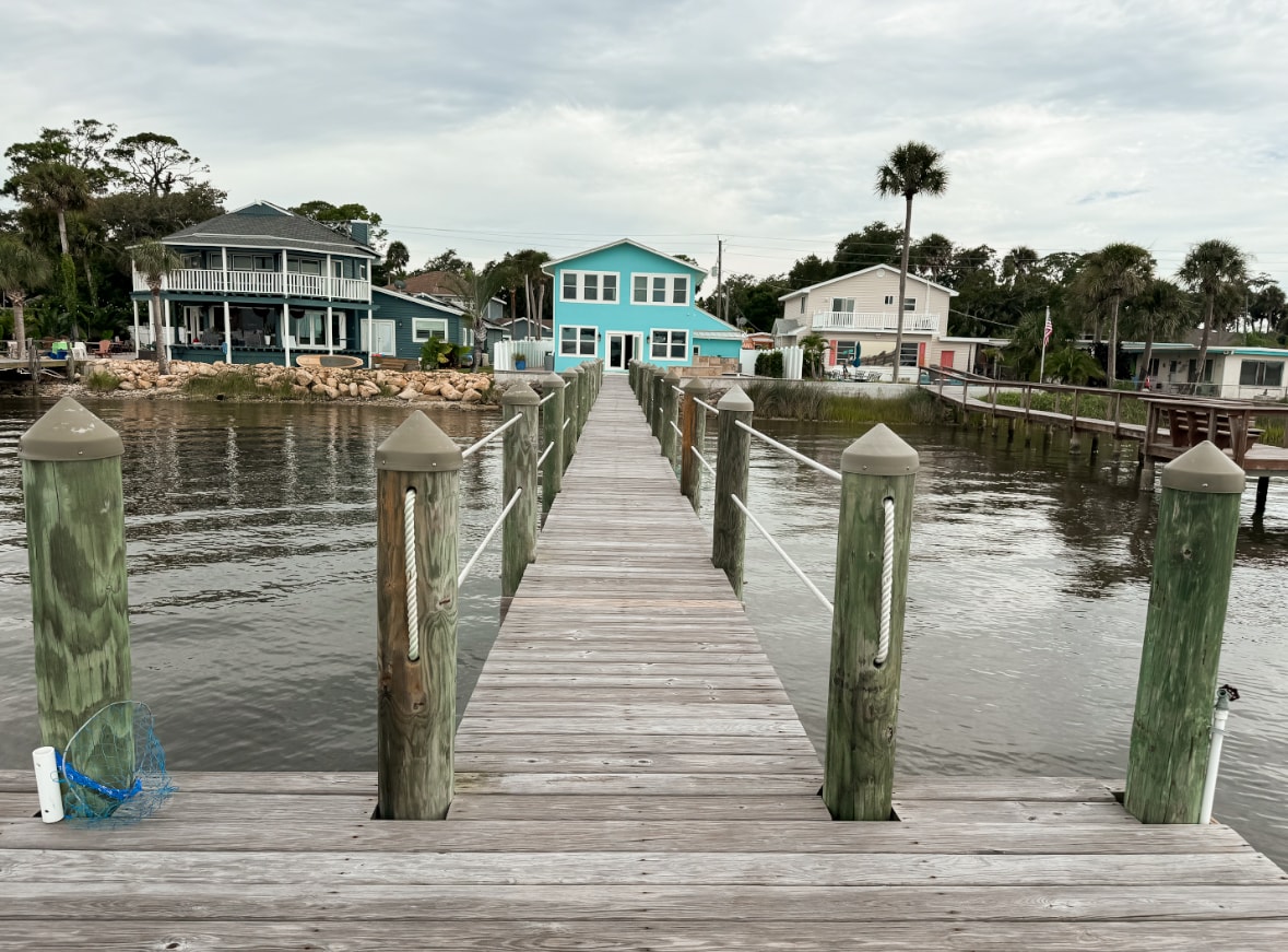 A wooden dock extends over calm water, leading towards a charming blue house. Tall wooden posts frame the dock, with a fishing net visible at the water’s edge. Lush greenery and various houses can be seen along the shoreline, creating a serene waterfront atmosphere.