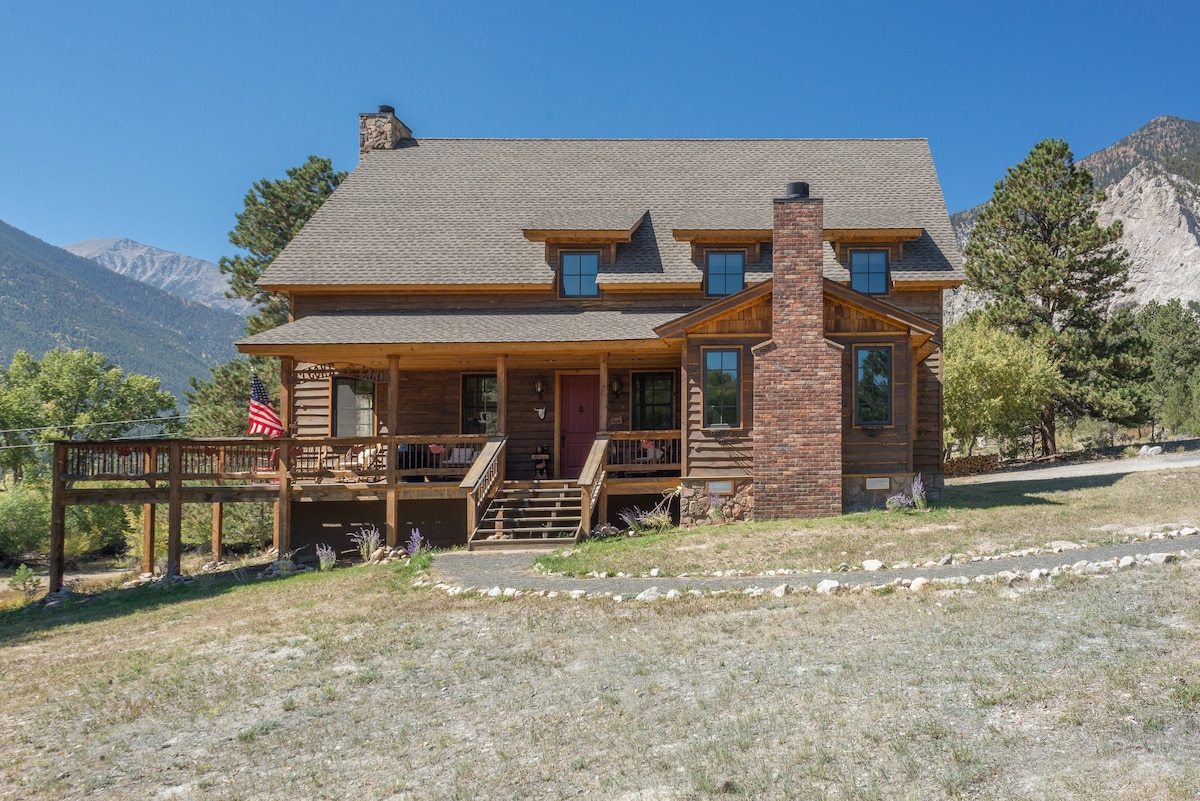 The exterior of a rustic wooden house is presented with a welcoming porch and a brick chimney, set against a backdrop of mountains. Surrounding trees and open space contribute to the serene environment. A deck extends from the side, adding to the outdoor living area.