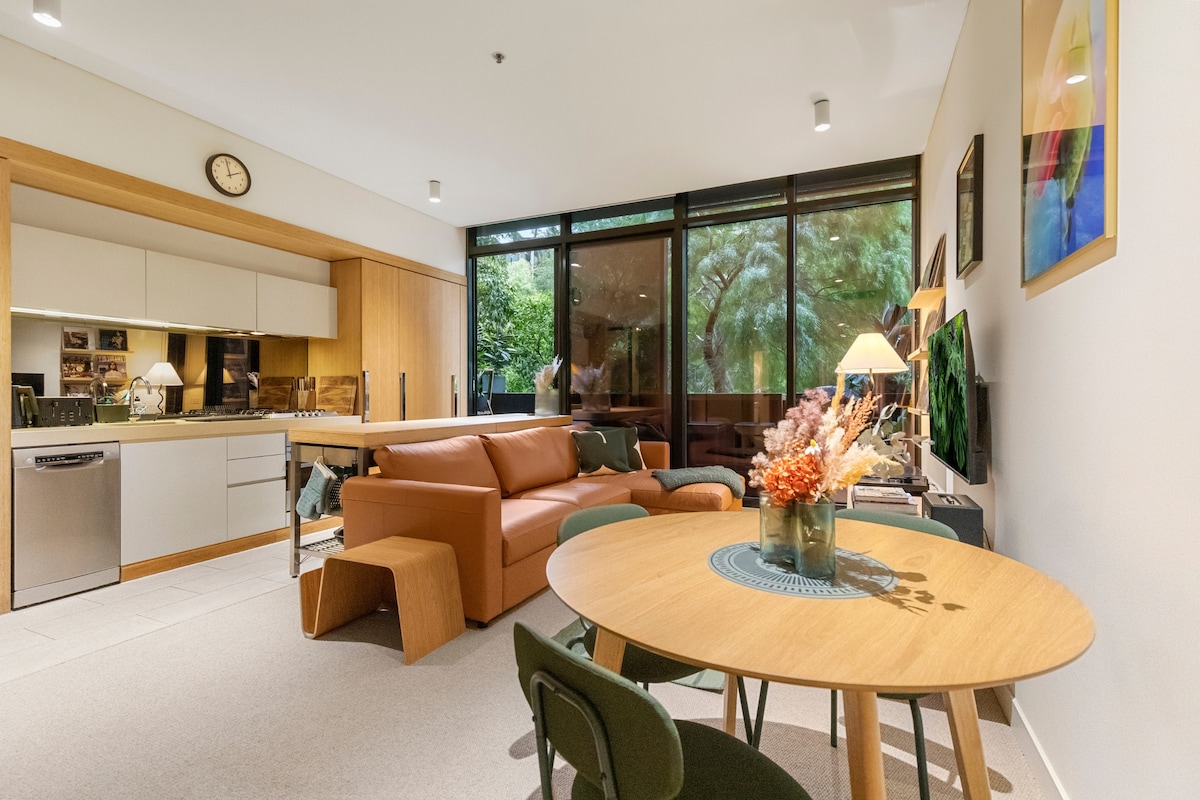 An open-plan living area showcases a comfortable brown couch paired with a wooden coffee table. A round dining table with green chairs is positioned nearby. Floor-to-ceiling windows provide ample natural light and a view of leafy surroundings, while a modern kitchen area can be seen in the background.