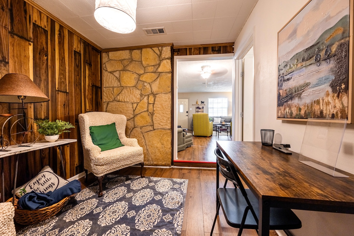 A cozy corner of the home is highlighted by a textured stone accent wall and wooden paneling. A comfortable armchair with a green cushion sits beside a small table with a decorative lamp. A desk is positioned nearby, providing a space for work or leisure.