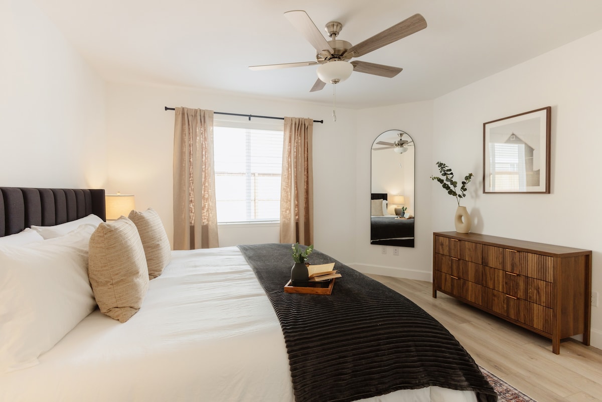 The primary bedroom features a spacious bed with plush bedding and decorative pillows. A dresser with a modern design is positioned against one wall, accompanied by a round mirror. Natural light filters through window treatments, enhancing the serene atmosphere.