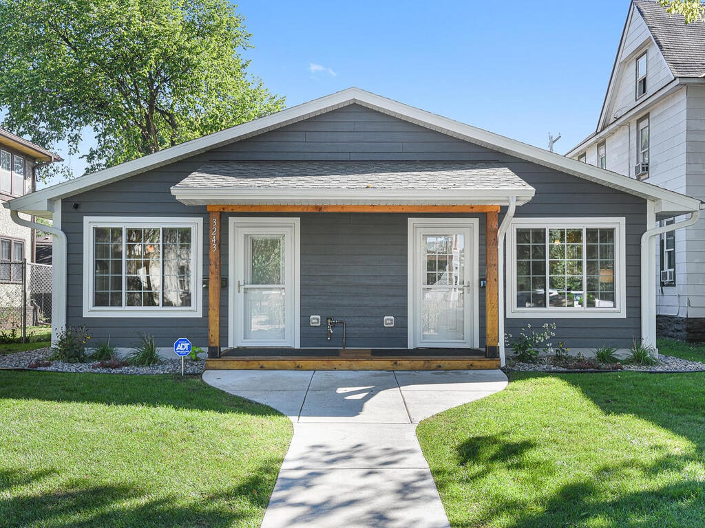 The front exterior of a charming single-story home is displayed, featuring a gray wooden facade with white trim. Two symmetrical entry doors are framed by large windows on either side. A well-maintained green lawn leads to a concrete pathway that guides guests toward the entrance.