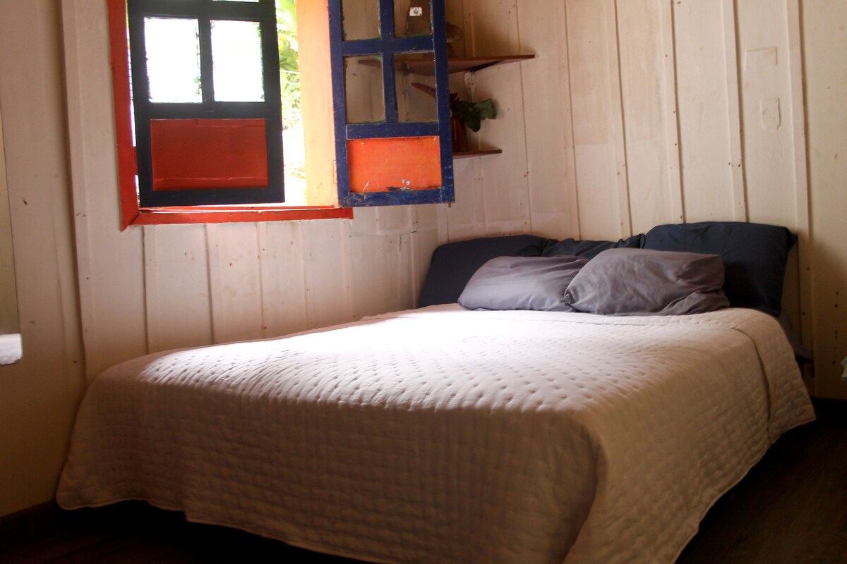 A cozy sleeping area features a spacious bed dressed in a light coverlet. Colorful shutters in orange and blue frame a window, allowing natural light to filter in. Wooden walls contribute to a warm ambiance, complemented by a shelf with greenery.