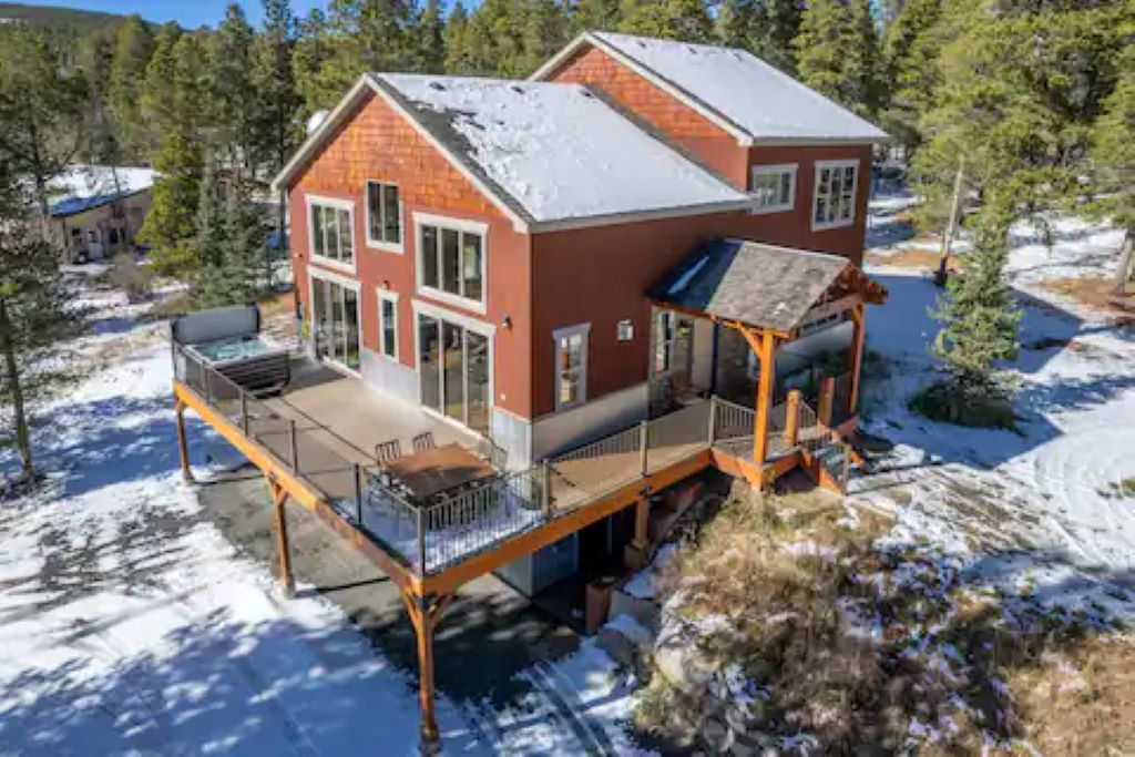 A spacious two-story home is displayed, featuring a warm red exterior and large glass windows. An expansive deck extends towards a snowy landscape, furnished with a dining set. Surrounding the house, evergreen trees provide a natural backdrop, enhancing the serene environment.