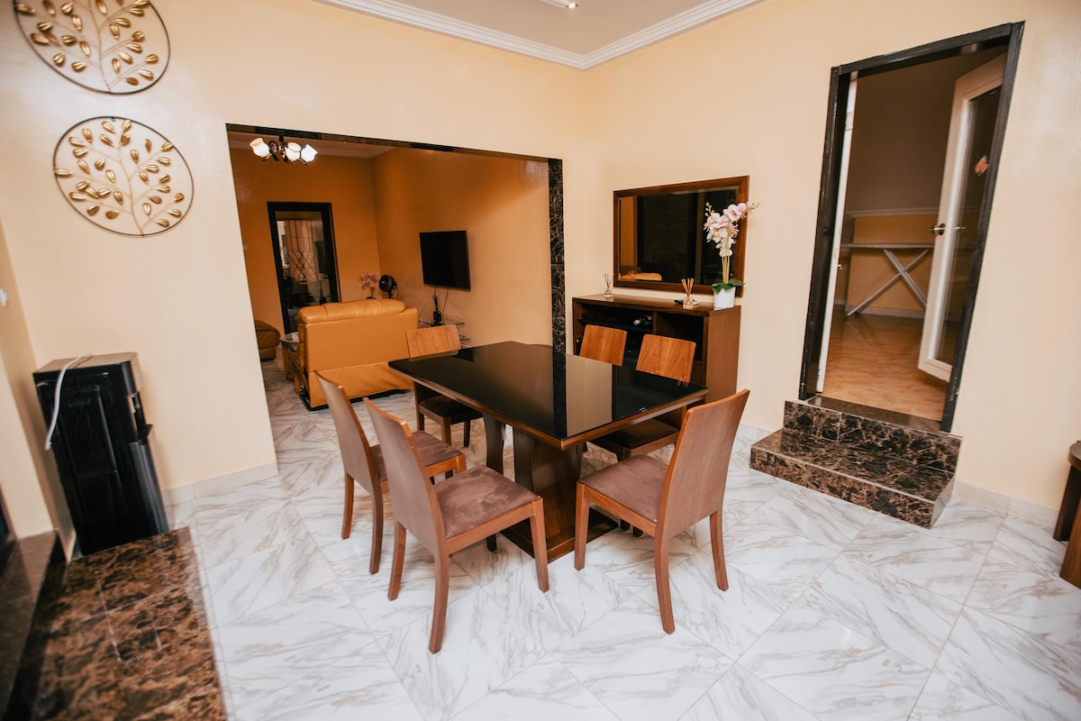 A dining area is presented with a sleek black table surrounded by six wooden chairs. The walls are painted in a warm tone, complemented by decorative elements, while a television is positioned on a side cabinet. Natural light enhances the space through an adjacent doorway.