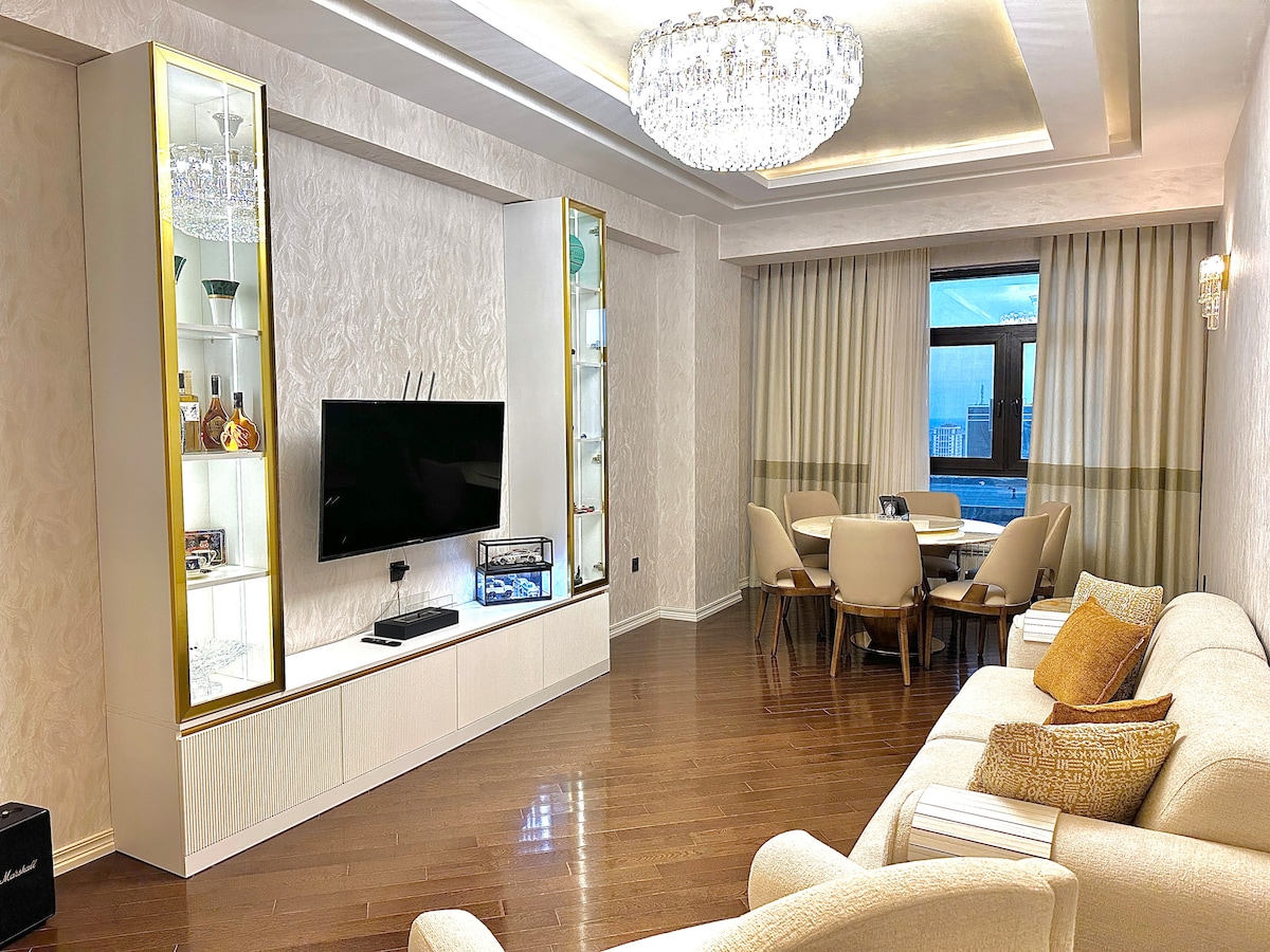 A stylish living area is presented with a comfortable sofa and a crystal chandelier above. A flat-screen TV is mounted on the wall, accompanied by a display cabinet showcasing decorative items. A dining space for four is visible near the window, adorned with soft curtains.