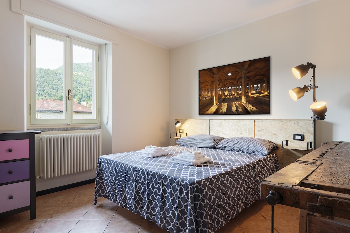 A well-lit bedroom features a spacious bed with patterned bedding and white linens. A large artwork adorns the wall above the bed. Two bedside lamps provide soft lighting, and a window offers a view of the mountainous landscape outside. A vintage dresser adds character to the room.