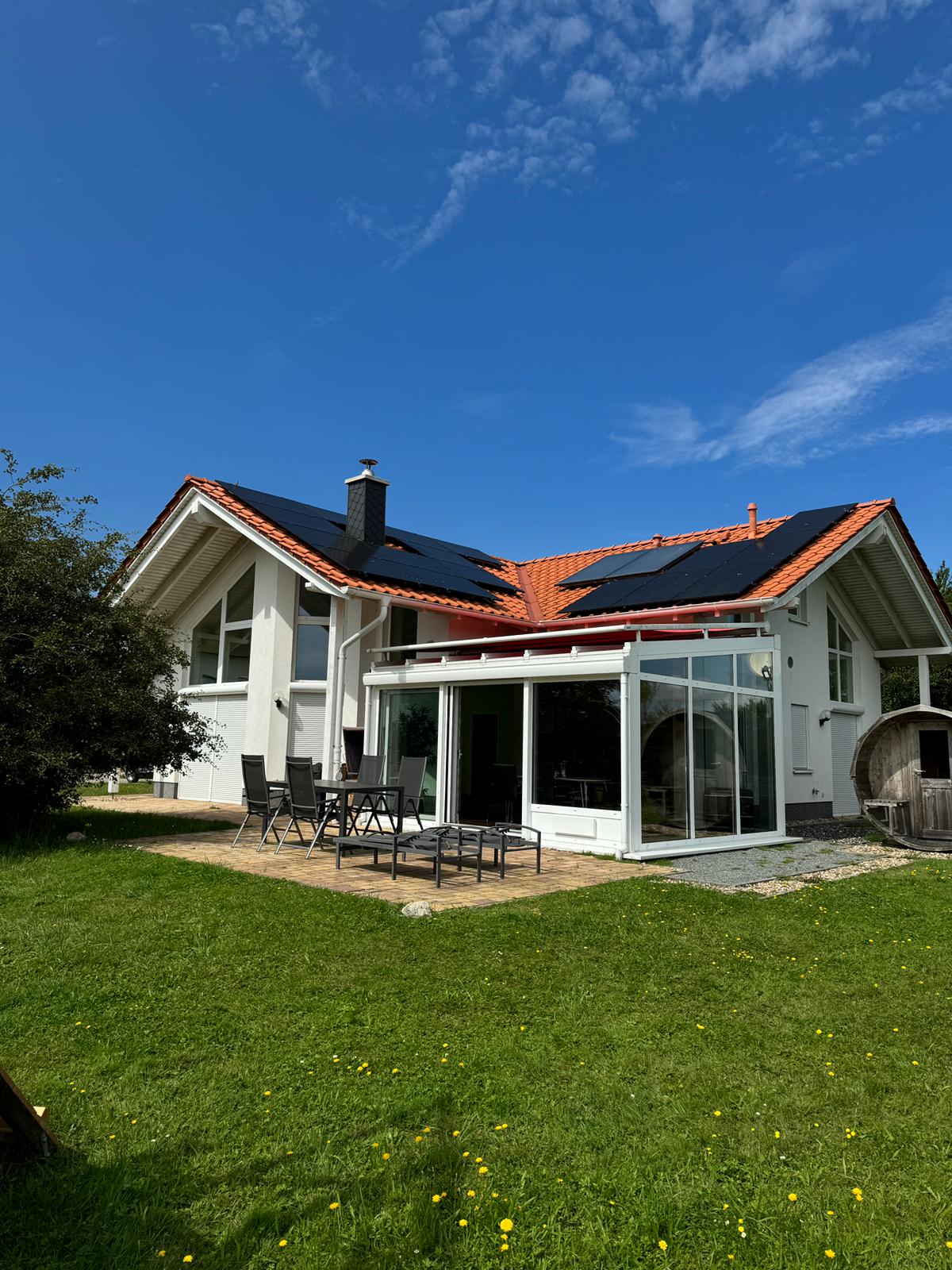 The exterior of a contemporary bungalow is shown, featuring a sloped roof with solar panels. A spacious patio is visible, furnished with a dining table and chairs, surrounded by a green lawn and trees. Clear blue skies provide a sunny backdrop.