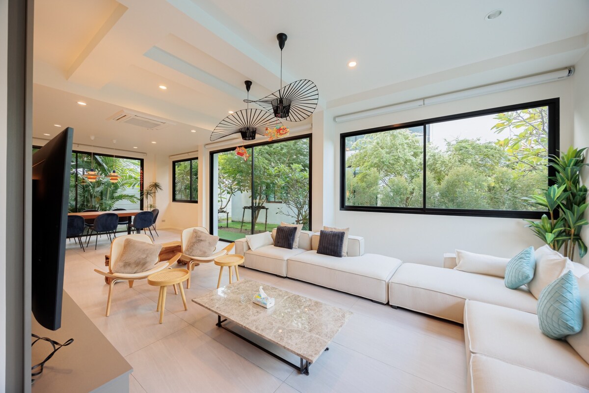 A modern living area features a spacious sectional sofa adorned with cushions, accompanied by two wooden chairs. Large windows allow natural light to fill the room, offering views of the greenery outside. A marble coffee table sits centrally, enhancing the clean, minimalist design.