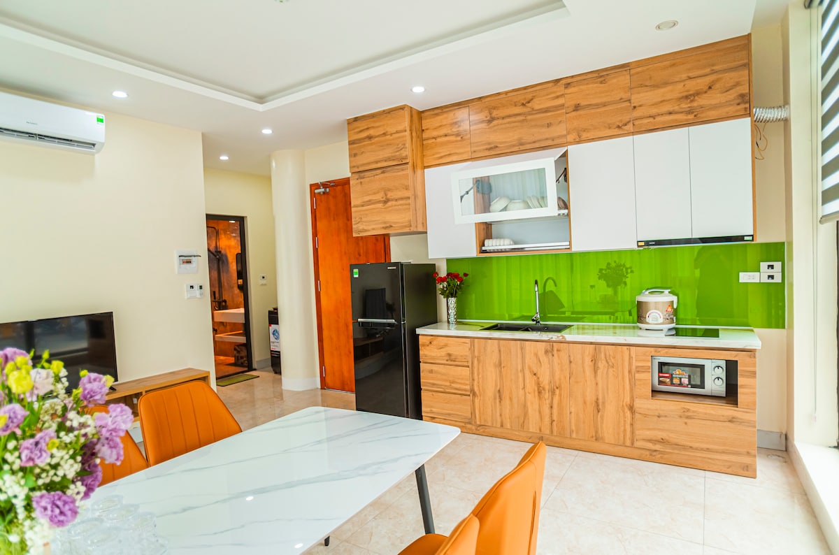 A modern kitchen is displayed with wooden cabinetry and sleek white countertops. Appliances include a refrigerator and stovetop, complemented by a vibrant green backsplash. A dining area features a marble table with orange chairs, arranged to create an inviting layout for meals.
