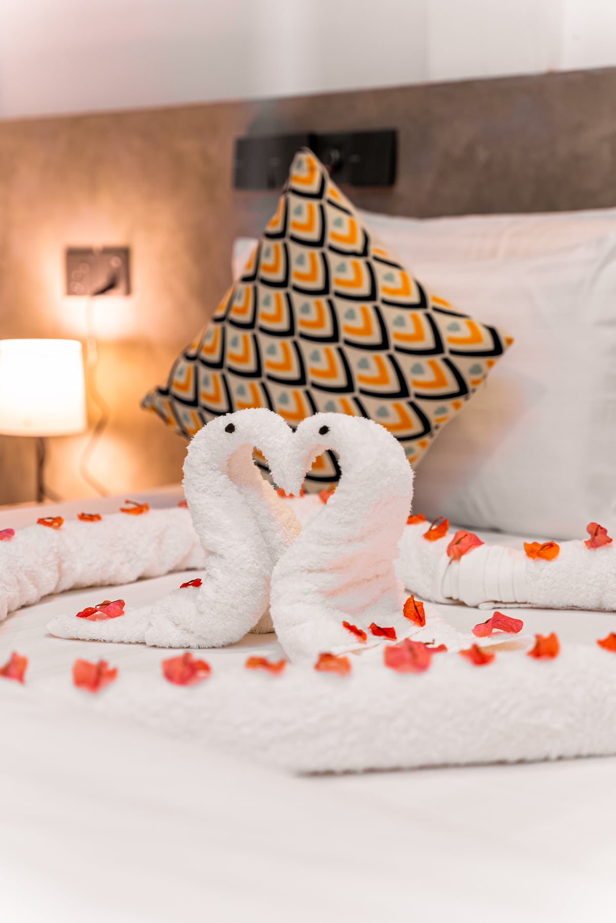 Two elegantly shaped towel swans are positioned on a bed, surrounded by petals in shades of red and orange. An accent pillow with a geometric pattern rests against the headboard, while soft lighting is provided by a bedside lamp nearby.