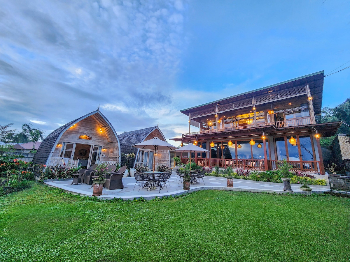 A spacious villa is surrounded by lush greenery, featuring a well-maintained lawn and outdoor seating arranged on a patio. The structure showcases a blend of traditional and modern architecture, with large windows and warm outdoor lighting creating an inviting atmosphere.